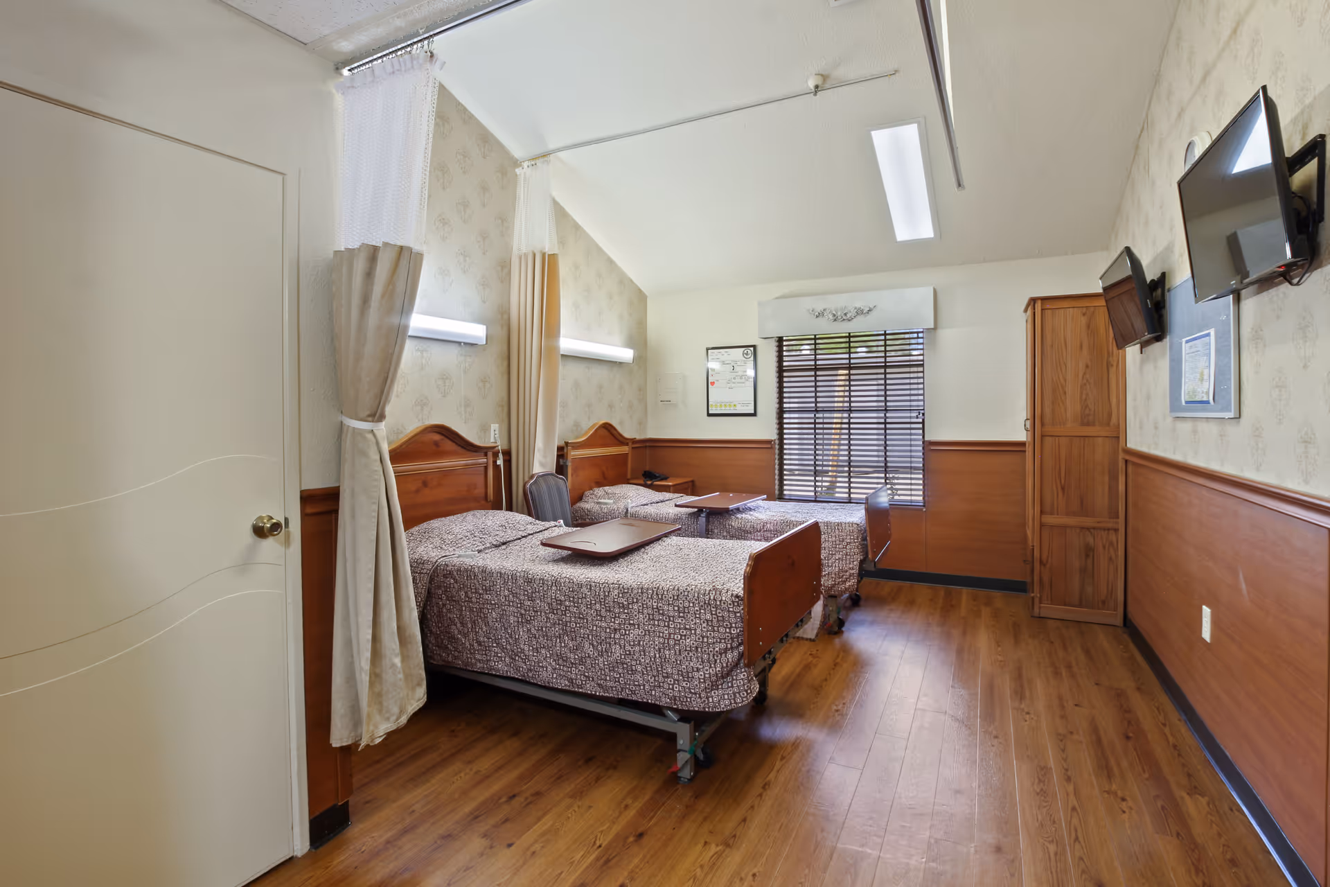 A shared nursing room with two hospital-style beds separated by a privacy curtain. Each bed has a patterned bedspread and an overbed table. The room features wood paneling on the lower walls, a window with blinds, two wall-mounted televisions, and a wooden cabinet. The floor is wood laminate, and the lighting is bright with ceiling fixtures and wall-mounted lights above each bed.
