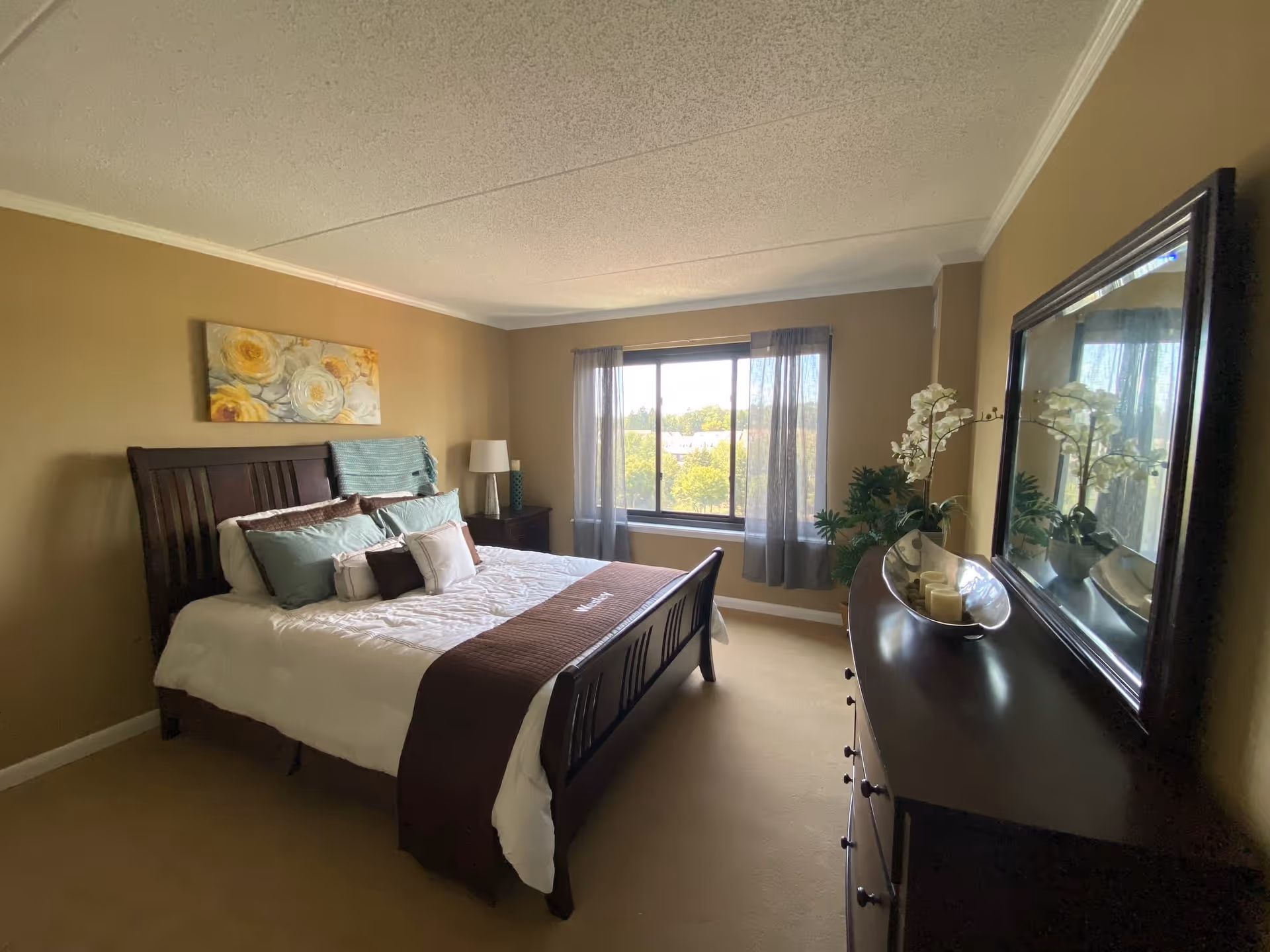 A cozy bedroom with a wooden bed frame, neatly made bed with white and brown bedding, multiple pillows, a nightstand with a lamp, a large window with sheer curtains letting in natural light, a dresser with a large mirror, and decorative plants and flowers.