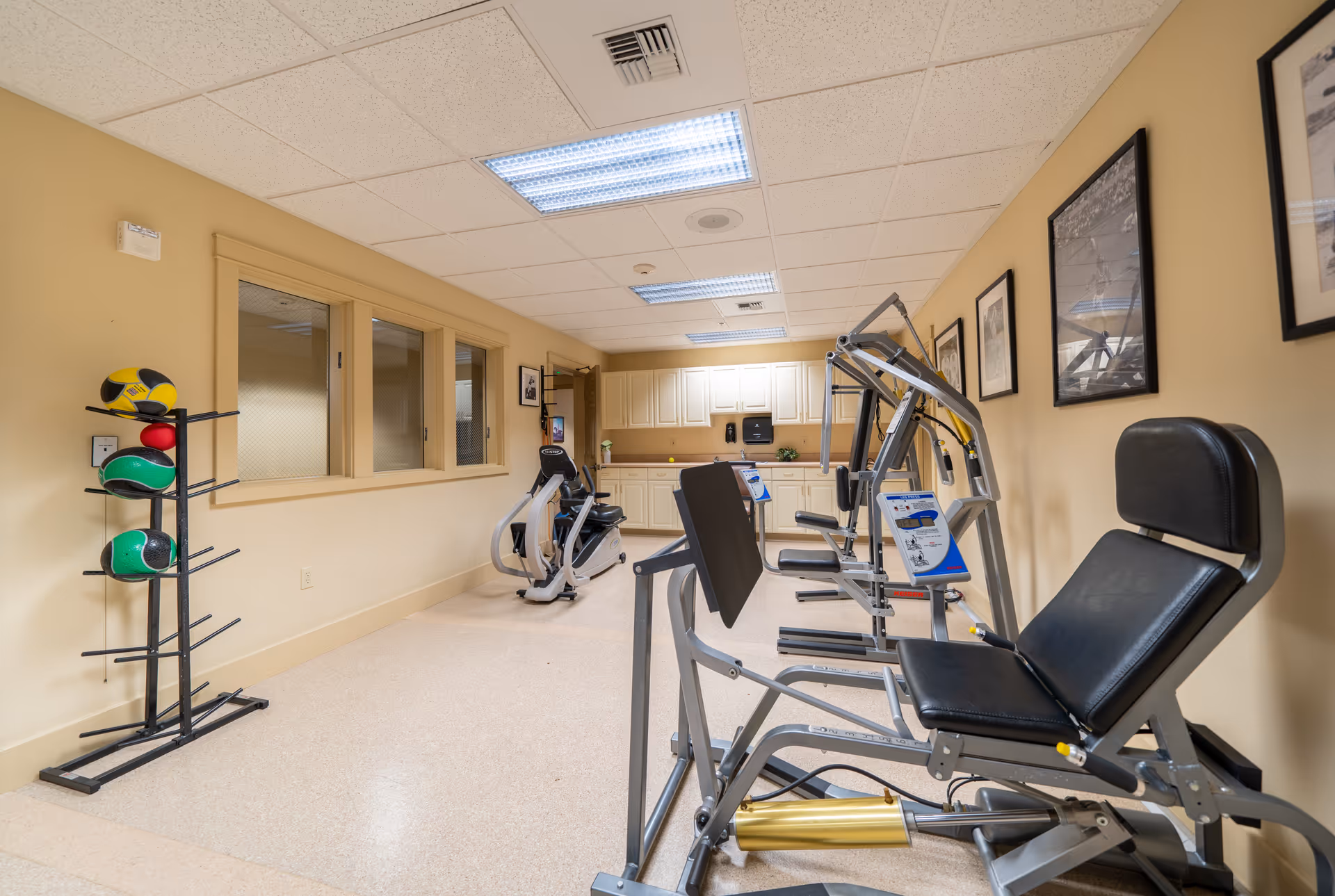 A fitness room in a senior living facility featuring exercise equipment including a seated leg press machine, a recumbent bike, and a rack holding several medicine balls. The room has beige walls, a tiled ceiling with fluorescent lights, and framed pictures on the wall. There is a counter with cabinets and a paper towel dispenser at the far end of the room.