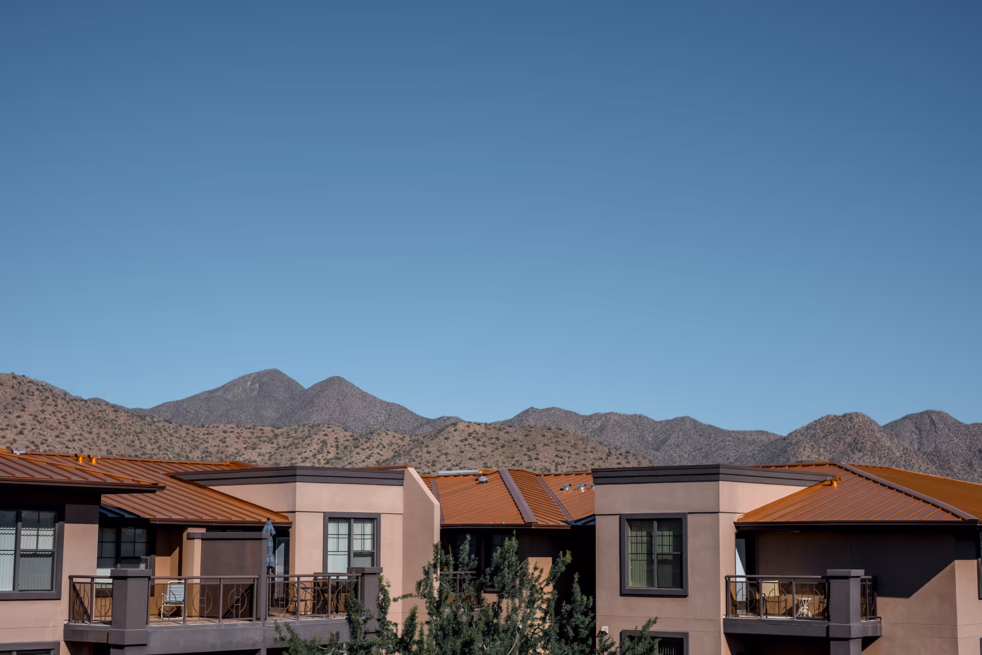 Exterior view of a residential building with balconies and reddish-brown roofs, set against a backdrop of rugged mountains under a clear blue sky.