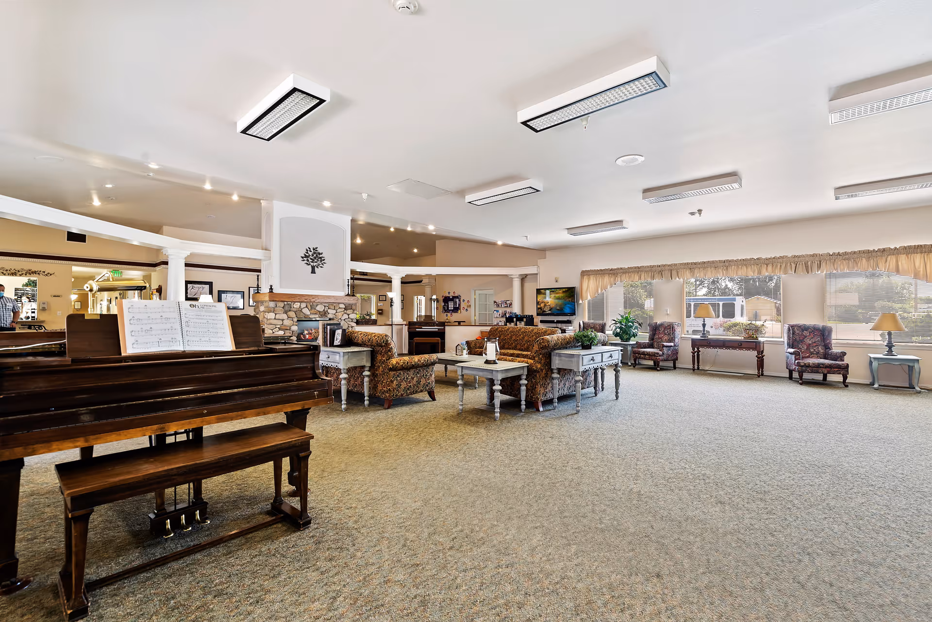 A spacious and well-lit assisted living facility common area featuring a wooden piano with sheet music, comfortable upholstered chairs and sofas arranged around coffee tables, a stone fireplace with a decorative tree artwork above it, large windows with beige curtains allowing natural light to fill the room, and various lamps and plants placed on side tables.