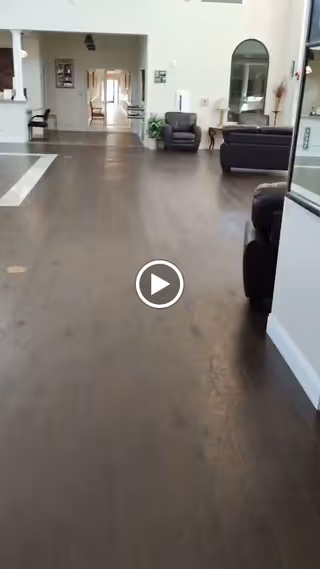 Interior view of a spacious assisted living facility hallway with dark wooden flooring, leather armchairs, a sofa, a large mirror on the wall, and a long corridor leading to other rooms.