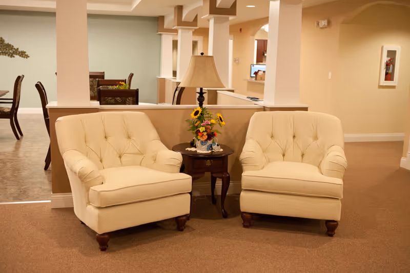 Two beige upholstered armchairs with wooden legs are placed on a carpeted floor with a small wooden side table between them. The table holds a lamp with a beige shade and a floral arrangement featuring sunflowers. In the background, there is a dining area with tables and chairs, and a light green wall with decorative elements.