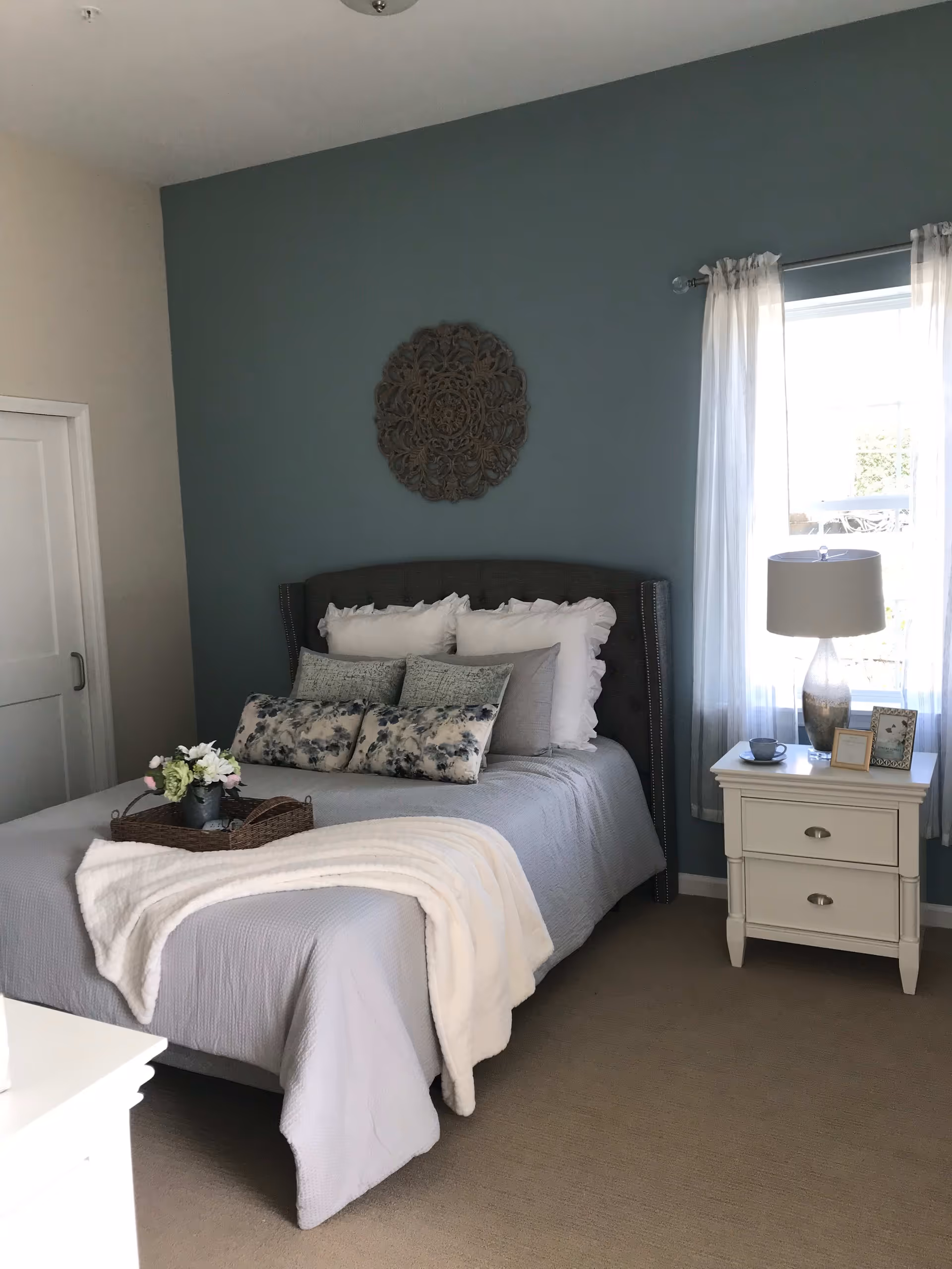A neatly made bed with multiple pillows and a white throw blanket in a bedroom. There is a decorative wall piece above the bed on a blue accent wall. A white nightstand with a lamp, framed photos, and a cup sits next to a window with sheer white curtains.