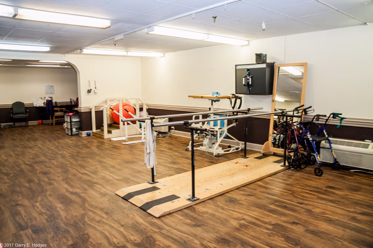A rehabilitation/therapy room with parallel walking bars, walkers, exercise equipment and chairs on a wood floor.