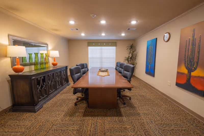 A well-lit conference room with a long wooden table surrounded by black leather chairs, a decorative sideboard with lamps and vases, and colorful cactus artwork on the walls.