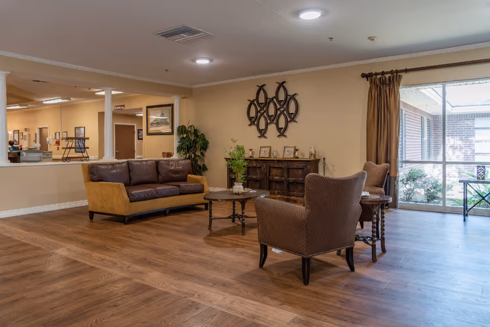 A spacious, warmly decorated lounge with a sofa, armchairs, coffee tables, and a large window looking out to a courtyard.