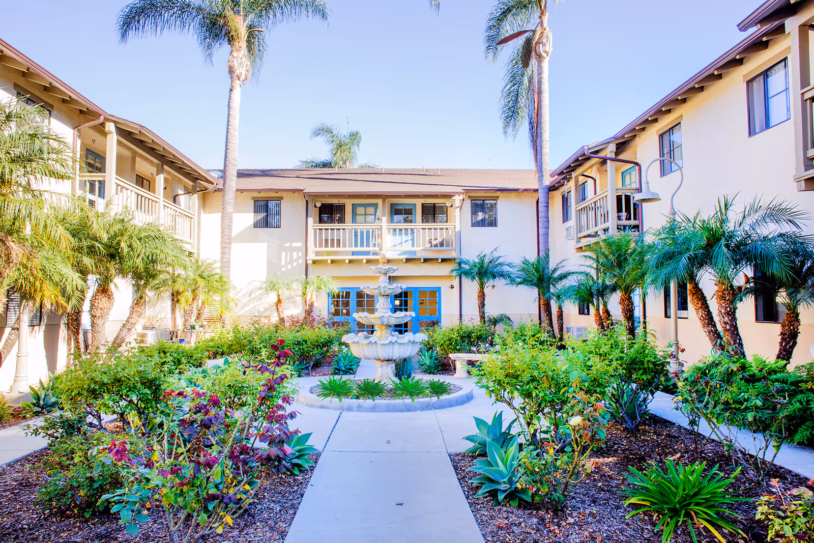 A bright courtyard garden at Ivy Park at San Juan Capistrano featuring a central multi-tiered fountain surrounded by lush greenery, palm trees, and flowering plants. The courtyard is enclosed by a two-story building with balconies and windows overlooking the garden.