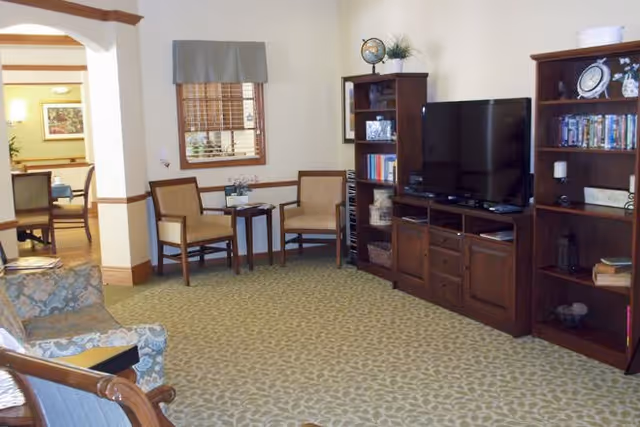 A cozy living room area with patterned carpet, two wooden chairs with cushions and a small table between them, a large flat-screen TV on a wooden entertainment center, and bookshelves filled with books and decorative items. There is a window with blinds and a valance, and an archway leading to another room with a dining table and chairs.