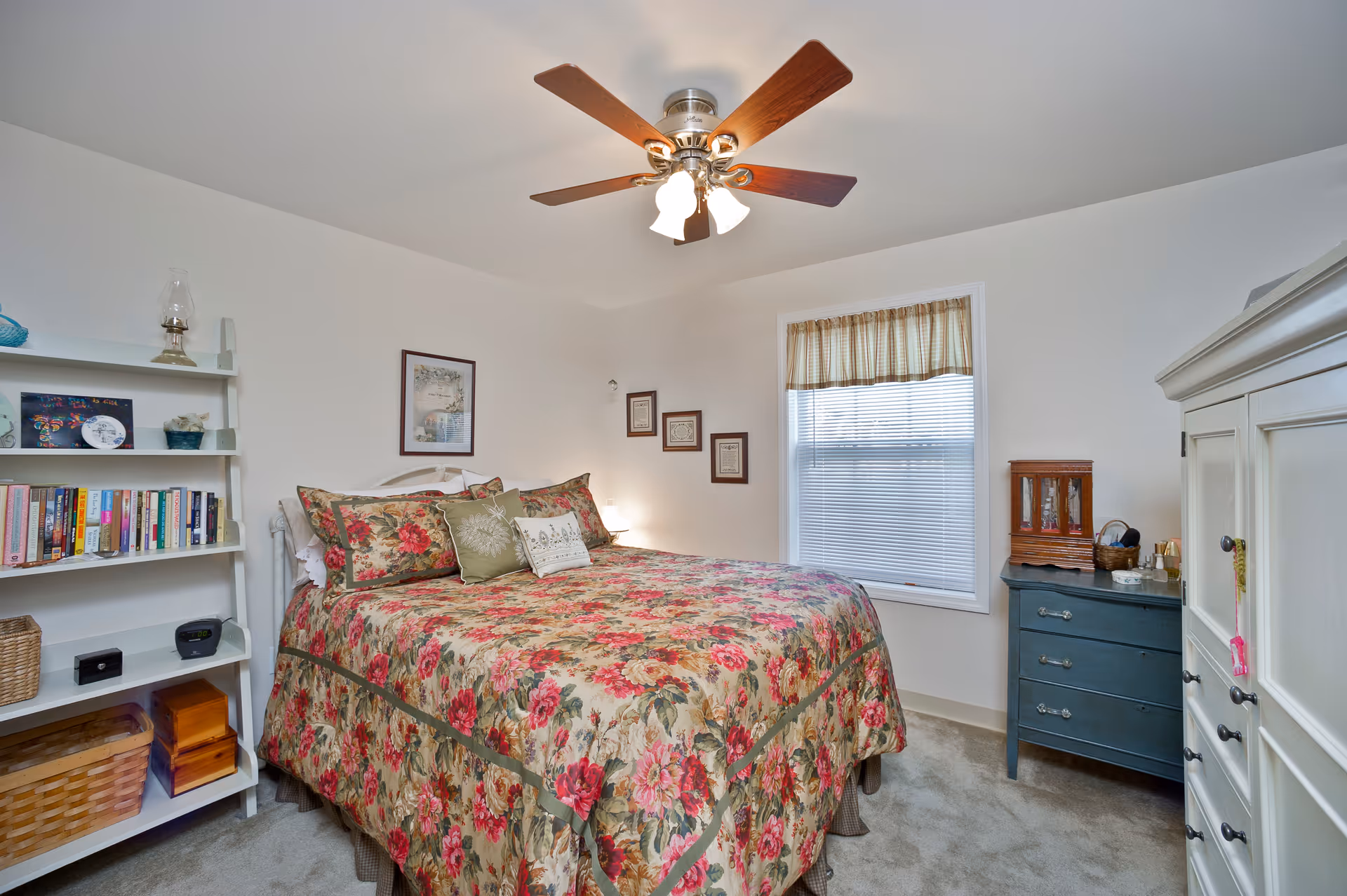 A cozy bedroom with a floral patterned bedspread on a bed, a ceiling fan with lights, a window with blinds and a valance, a white bookshelf filled with books and decorative items, a blue dresser with a wooden jewelry box and other small items, and a white wardrobe.