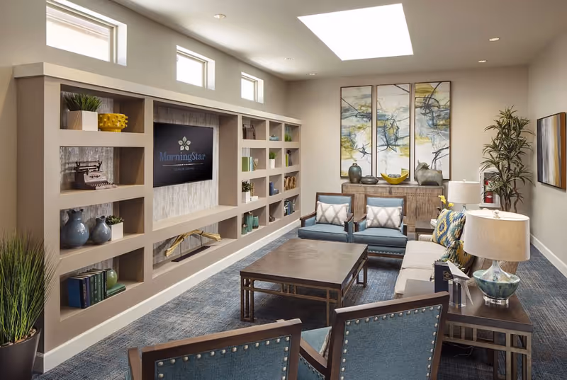 A modern living room in an assisted living facility featuring a large built-in shelving unit with decorative items and a mounted TV displaying the MorningStar logo. The room has a blue carpet, a coffee table, two blue armchairs, a beige sofa with patterned pillows, side tables with lamps, abstract wall art, and a tall potted plant in the corner.