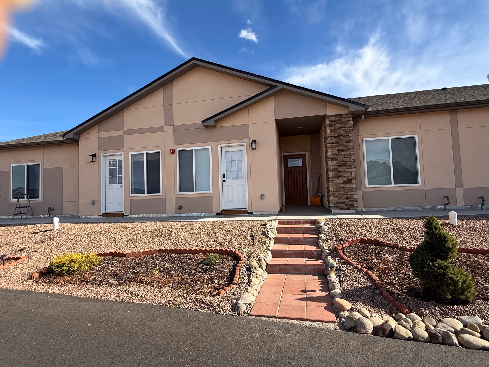 Exterior view of a single-story building with beige walls and a dark roof under a blue sky with some clouds. The building has multiple windows and white doors, with a small set of tiled steps leading up to a covered entrance supported by a stone pillar. The front yard features landscaped areas with rocks, mulch, small bushes, and a paved walkway.