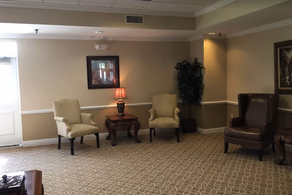 A quiet sitting area in a senior living facility with two beige upholstered armchairs and one dark brown leather armchair arranged around a wooden side table with a lamp. The walls are painted beige with white trim, and there are framed paintings and a potted plant in the corner. The floor is carpeted with a patterned design.