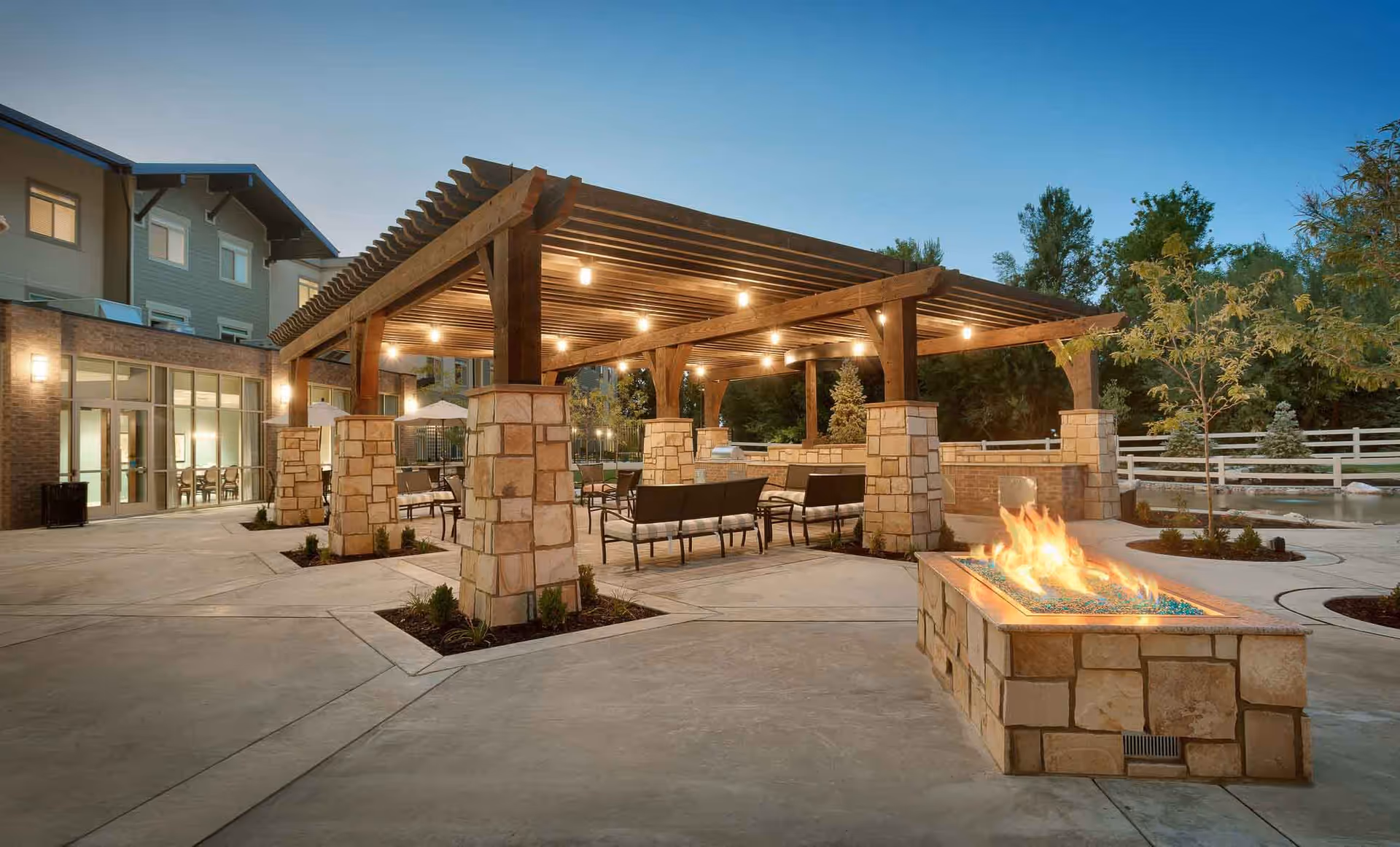 Outdoor patio area at Creekside Assisted & Senior Living featuring a large wooden pergola with string lights, stone pillars, seating arrangements with chairs and benches, and a rectangular stone fire pit with flames. The background includes part of the building and trees under a clear evening sky.