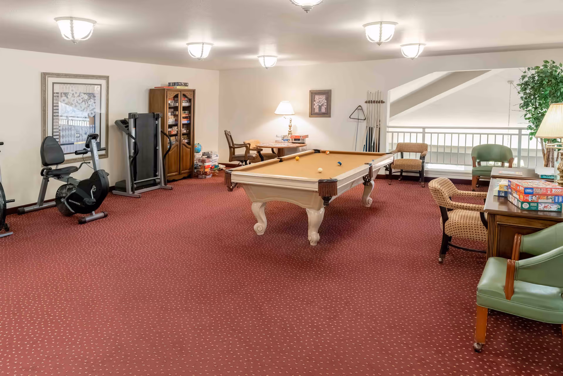 A recreational room with a pool table in the center, exercise equipment including a stationary bike and treadmill on the left, a cabinet filled with board games, and several chairs and tables around the room. The room has red carpet flooring, beige walls, and ceiling lights.