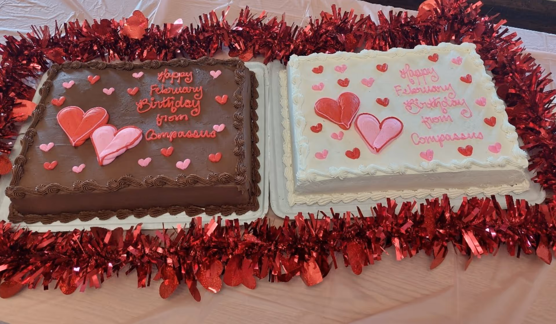 Two decorated sheet cakes—one chocolate and one white—adorned with pink and red hearts and surrounded by red tinsel on a table.