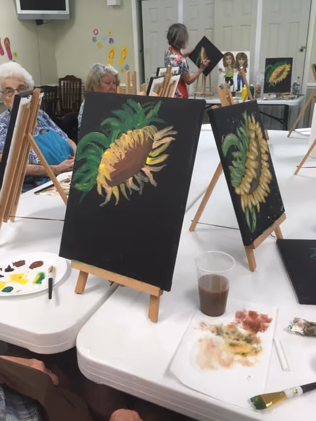 Several elderly individuals seated around tables engaged in a painting activity. Easels hold canvases with sunflower paintings in progress. Paint palettes, brushes, and a cup with brown liquid are on the tables. The room has light-colored walls with some decorations and closed white doors in the background.