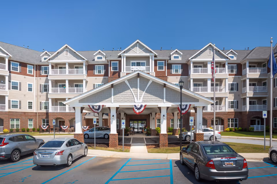 Front exterior view of a multi-story senior living facility named Harmony at Five Forks, featuring a covered entrance with white columns and patriotic bunting. Several cars are parked in the parking lot in front of the building under a clear blue sky.