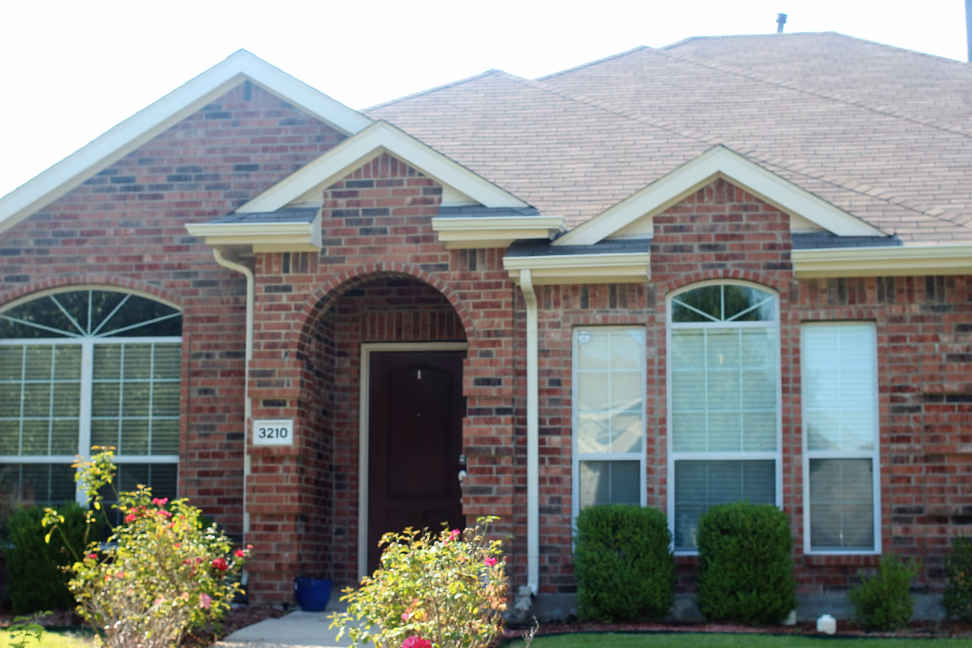 Front exterior view of a single-story brick residential building with a dark brown front door under an arched entryway, two large windows with white blinds, and neatly trimmed bushes and flowering plants in the front yard.