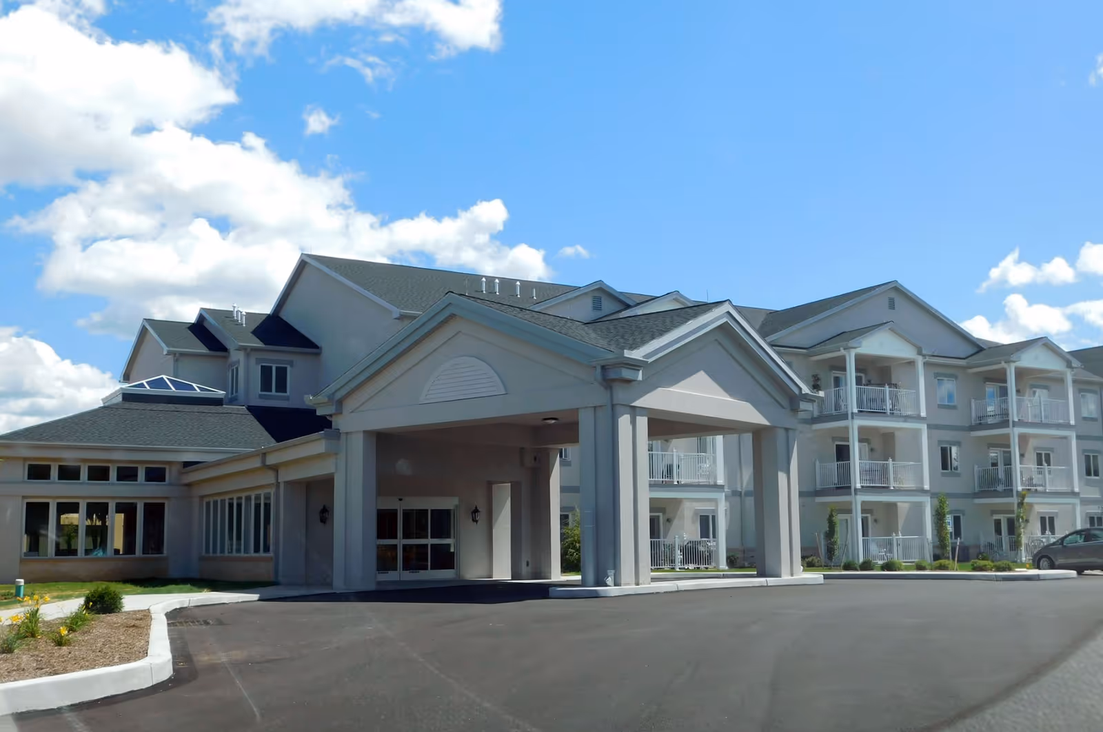 Exterior view of Londonderry Village senior living facility showing a large building with multiple balconies, a covered entrance, and a clear blue sky with some clouds.