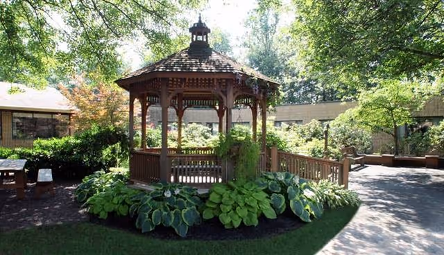 A wooden gazebo surrounded by lush green plants and bushes in a garden area with trees and a paved walkway, located within the grounds of Luther Woods Nursing & Rehabilitation Center.