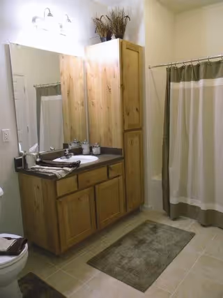 A bathroom with a wooden vanity featuring a sink and countertop, a large mirror above it, and a tall wooden cabinet next to the vanity. There is a shower with a beige and brown curtain, a toilet partially visible on the left, and two brown bath mats on the tiled floor. The walls are light-colored and there is a light fixture above the mirror.