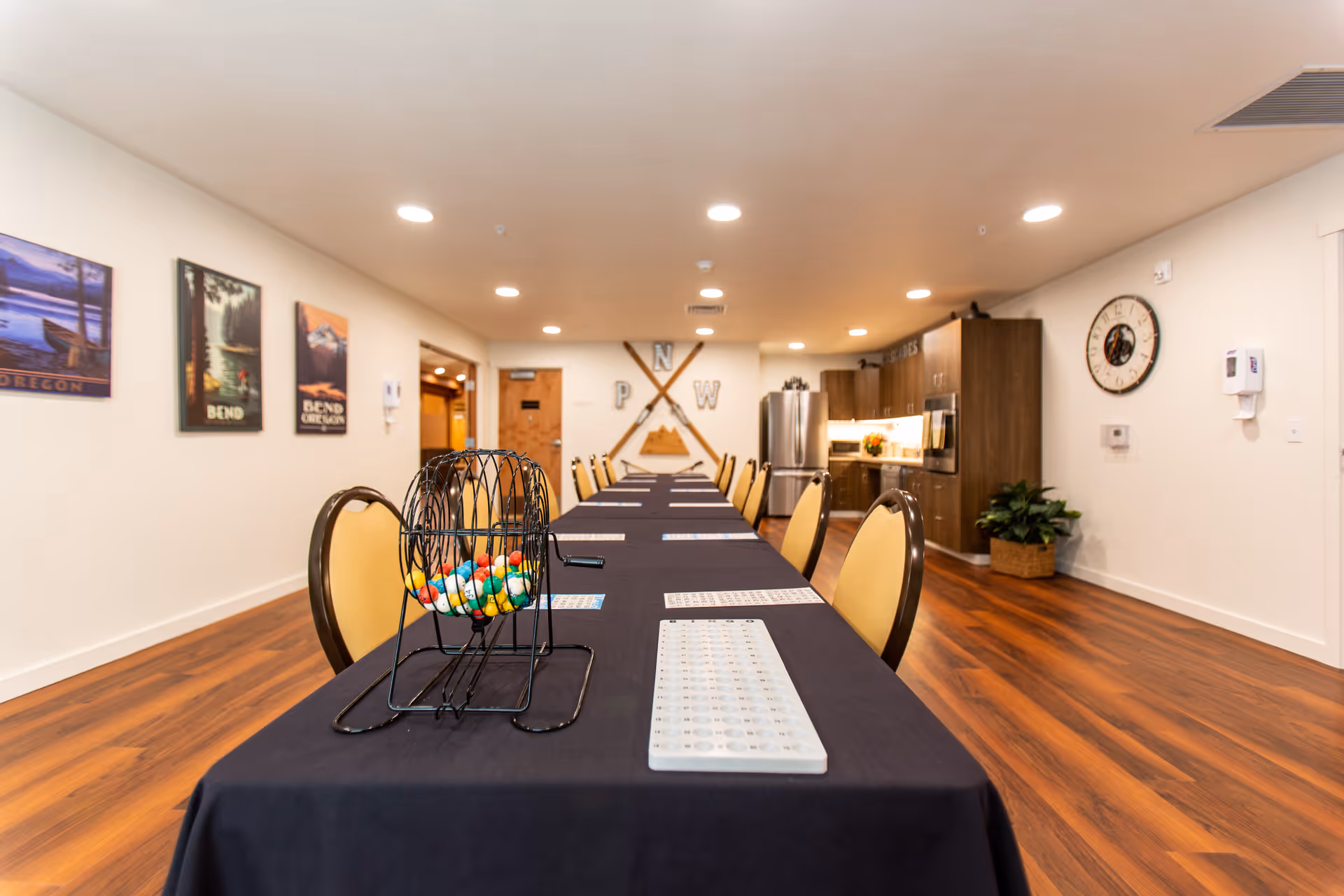 Long community activity room with a black-covered table set for bingo, chairs along both sides, and a kitchenette at the far end.