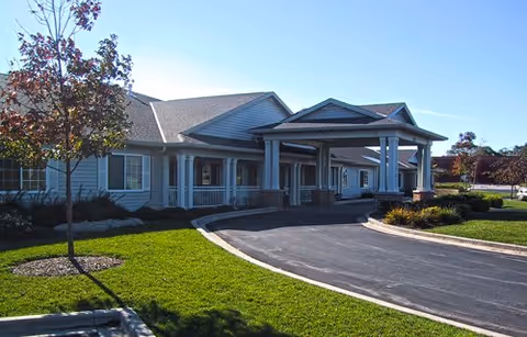 Exterior view of a single-story assisted living facility with a covered entrance, a paved driveway, green lawn, and a small tree in front under a clear blue sky.