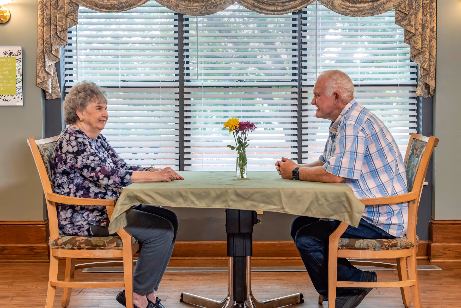 Two elderly people sit across from each other at a small table with a vase of flowers in front of a large window with blinds.