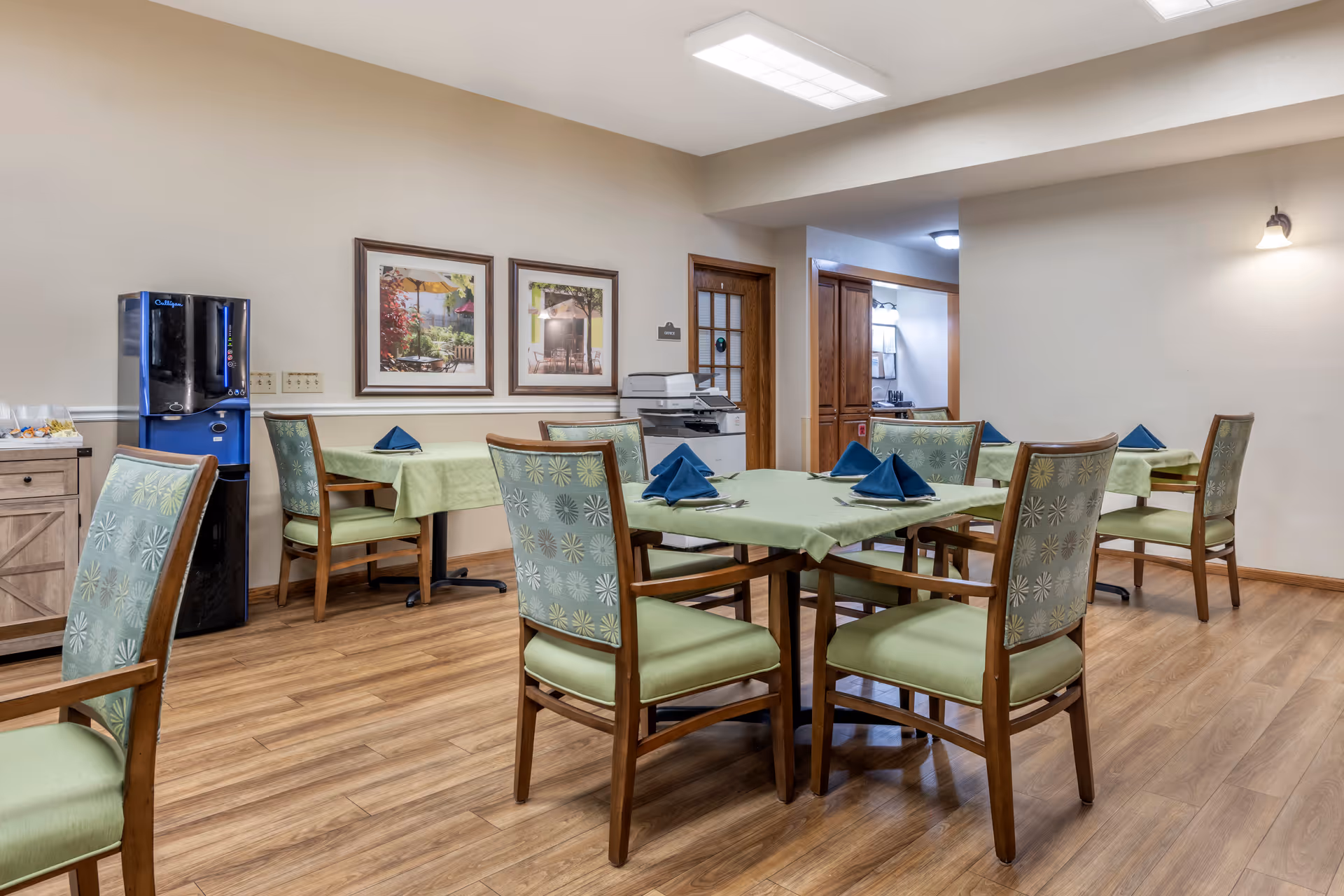 A dining room in a senior living facility with wooden floors, several tables covered with light green tablecloths, and chairs with green patterned upholstery. Each table has neatly folded blue napkins and silverware. The room has beige walls with two framed pictures and a water dispenser in the corner.