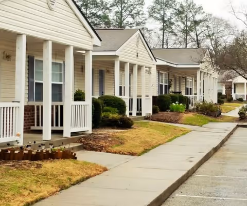 Row of single-story apartment units with small covered porches, white railings, and beige siding, bordered by a sidewalk and landscaped with bushes and grass under an overcast sky.