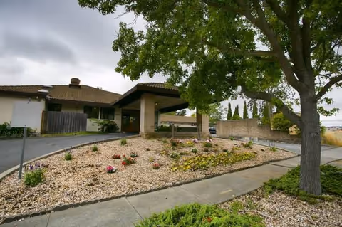 Entrance to a single-story senior facility with a stone garden, driveway, and large tree.