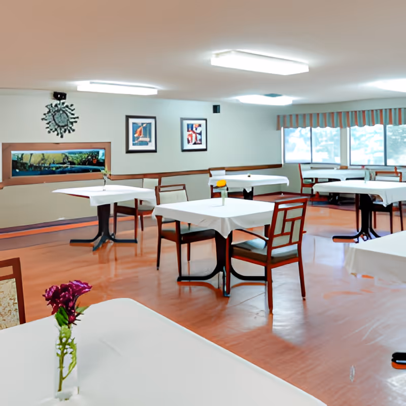 Dining room with multiple tables covered in white tablecloths, wooden chairs, small vases of flowers, and large windows.