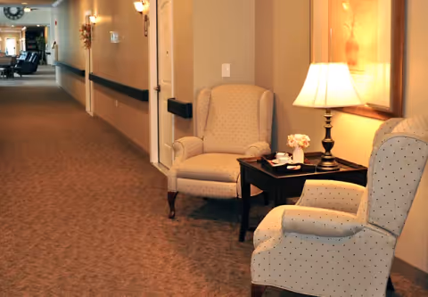 A well-lit hallway in a senior living facility with two upholstered armchairs and a small wooden side table with a lamp and a small flower arrangement. The hallway has carpeted flooring, beige walls, and wall-mounted light fixtures. Doors and handrails line the corridor.