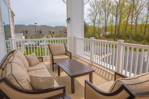 Outdoor balcony seating area with cushioned chairs and a coffee table, overlooking a parking lot and trees in the background.
