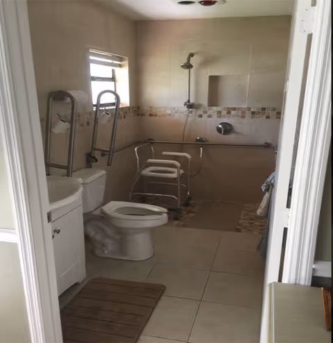 Accessible bathroom with a toilet and sink, grab bars, and a tiled walk‑in shower area with a shower chair.