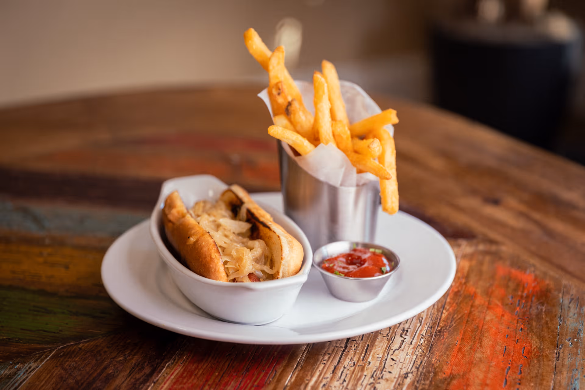 A plate with a hot dog topped with sauerkraut in a white dish, a metal cup filled with French fries, and a small metal container of ketchup on a rustic wooden table.