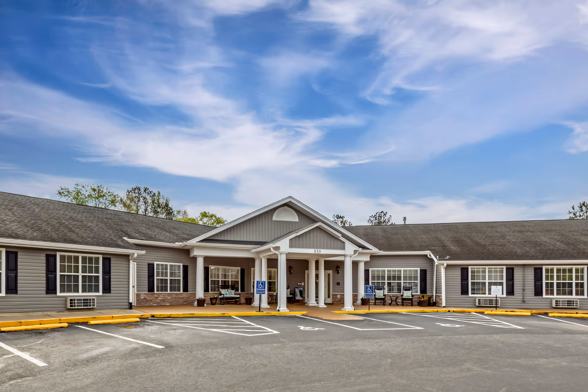 Front exterior view of Brookdale Smithfield facility showing a single-story building with gray siding, white columns at the entrance, several windows with black shutters, and a parking lot with designated handicapped parking spaces under a partly cloudy blue sky.