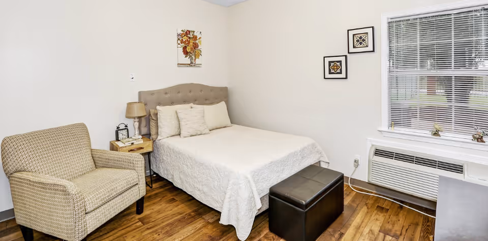 A simple bedroom with a bed, upholstered armchair, nightstand and lamp, window with blinds and an air-conditioning unit.