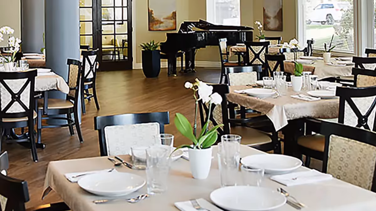 A dining room with tables set with white plates, glasses, and silverware. Each table has a small white potted plant with white flowers. The room has wooden floors, beige walls, and large windows letting in natural light. In the background, there is a black grand piano and some framed artwork on the walls.