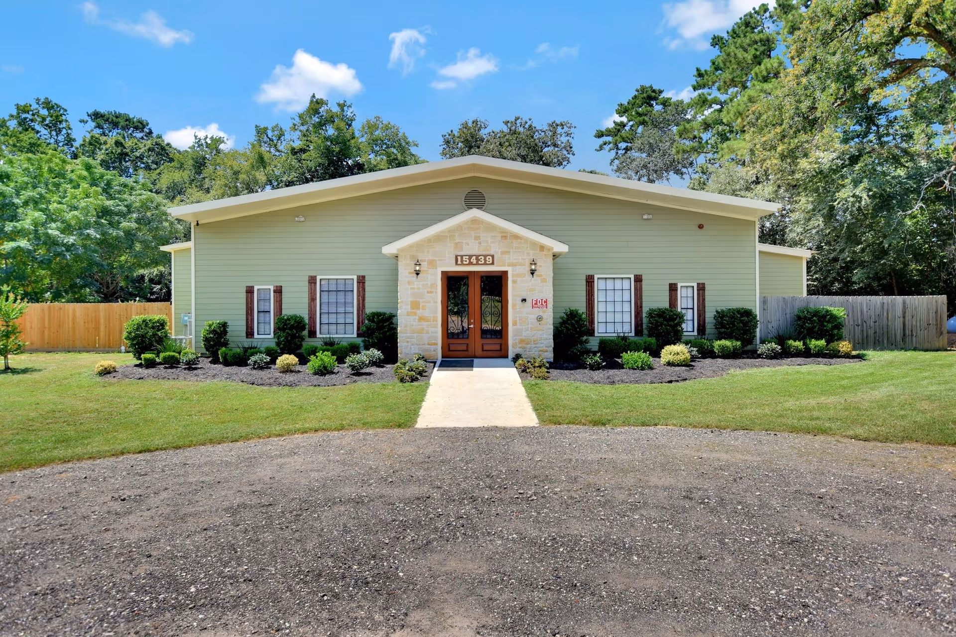 Front exterior view of a single-story building with light green siding and a stone entrance. The building has several windows with brown shutters and is surrounded by neatly trimmed bushes and a green lawn. A concrete walkway leads to the double wooden front doors under a small gabled roof. Trees and a wooden fence are visible in the background under a blue sky with a few clouds.