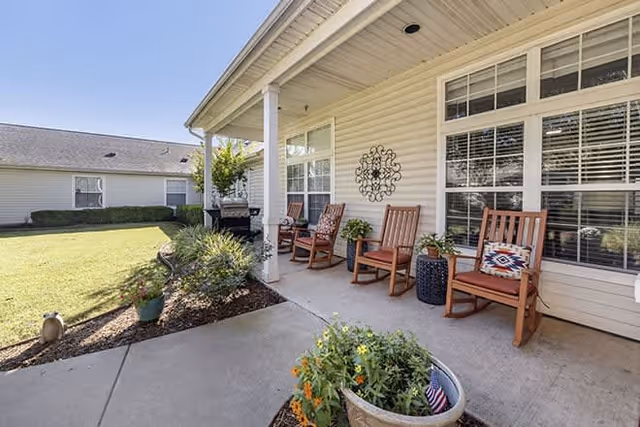 Covered patio area at Brookdale Stillwater with four wooden rocking chairs, potted plants, and decorative wall art. The patio overlooks a well-maintained lawn and garden area with shrubs and flowers.