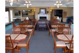 A spacious dining room with multiple wooden tables and chairs arranged neatly. The room features a carpeted floor, a fireplace at the far end, and several ceiling light fixtures. There are windows on the left side allowing natural light to enter.