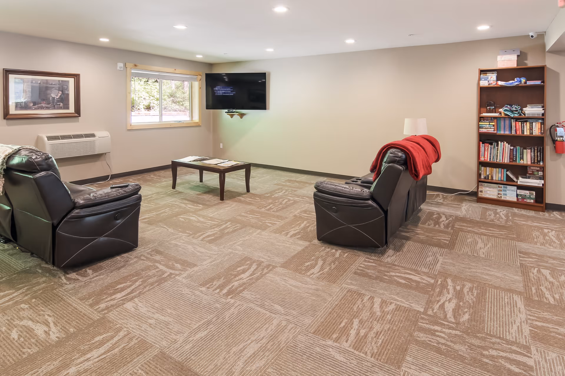 A cozy living room with two black leather recliners facing a wall-mounted flat-screen TV. A wooden coffee table with magazines is placed between the chairs. There is a window with a wooden frame letting in natural light, a framed picture on the wall, and a tall bookshelf filled with books and games. The floor is carpeted with a patterned design, and a red blanket is draped over one recliner.