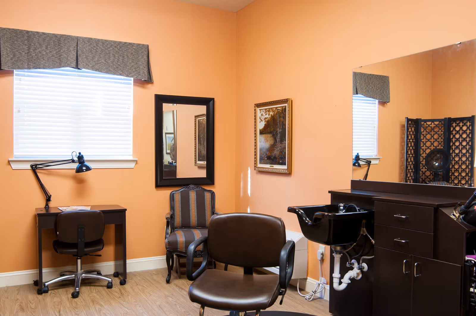 A room with peach-colored walls featuring a black salon chair in front of a black hair washing sink and cabinet. There is a window with blinds and a gray valance, a small desk with a black lamp and chair, a striped upholstered armchair, a framed painting on the wall, and a large mirror reflecting part of the room.