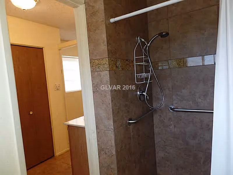 Tiled walk-in shower with a handheld showerhead, grab bar and curtain beside a bathroom vanity and hallway.