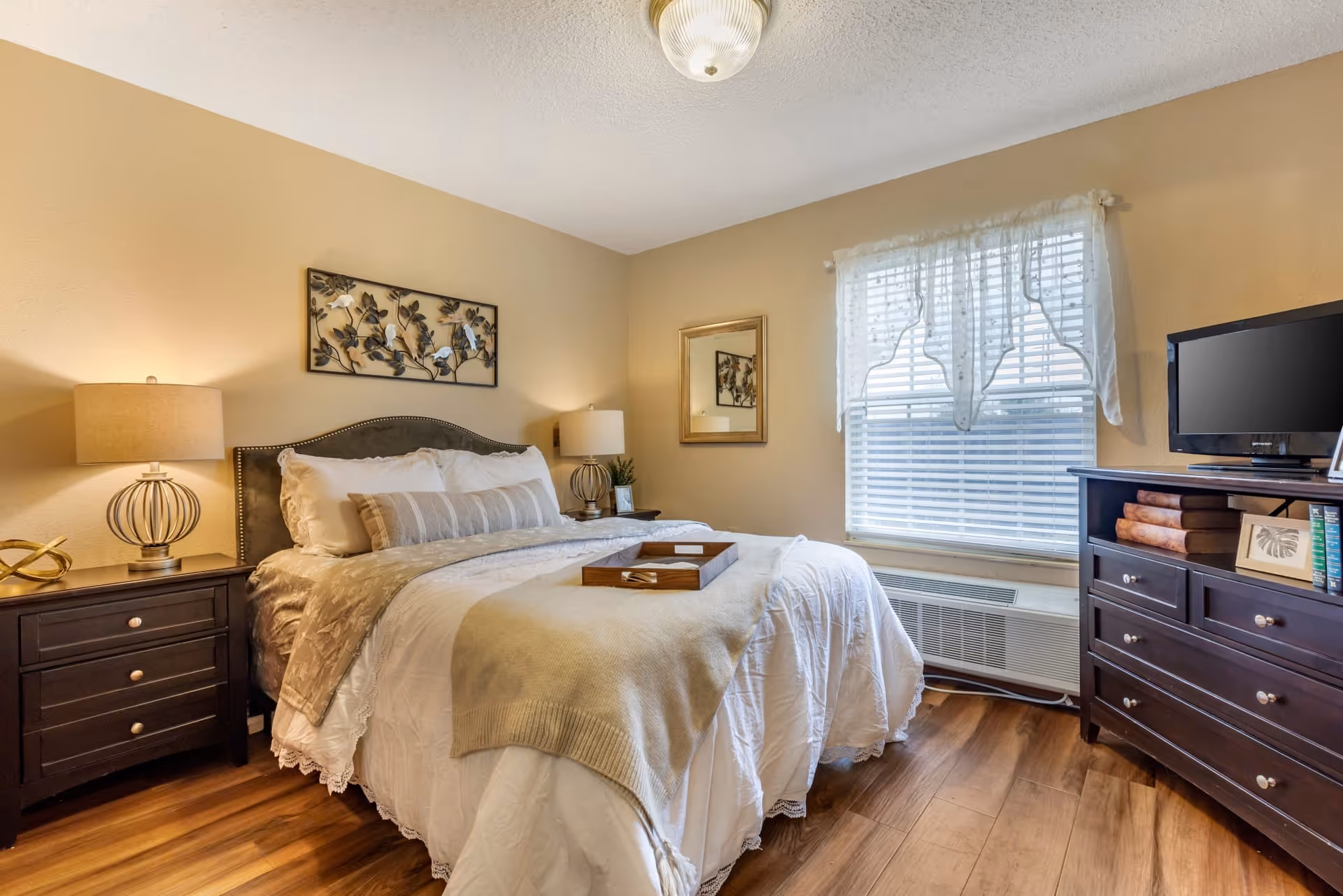 A cozy bedroom with a neatly made bed featuring white and beige bedding, two bedside tables with lamps, a decorative wall art above the bed, a window with sheer curtains, and a dark wooden dresser with a small TV on top.