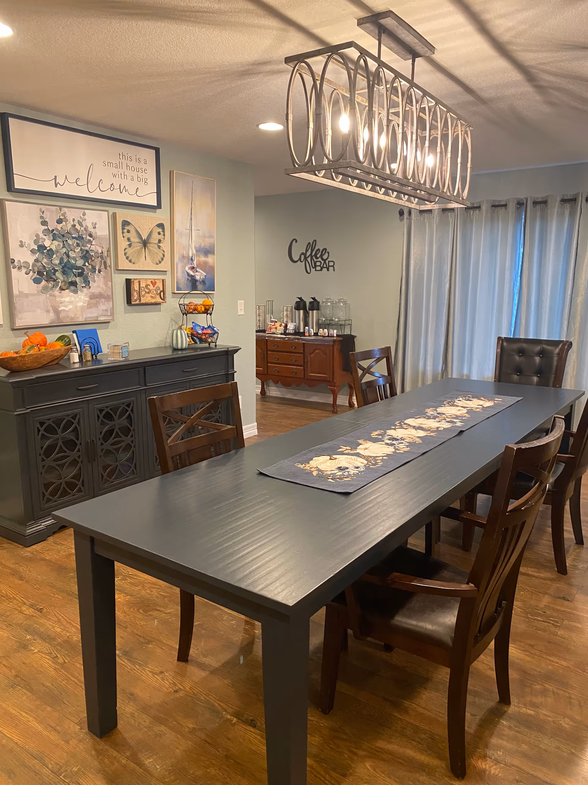 A dining room with a long black table and six wooden chairs. On the table is a decorative runner with a floral pattern. The room has wooden flooring and light-colored walls adorned with framed artwork, including a butterfly and a sailboat. A sideboard with a bowl of fruit and other items is against one wall. In the background, there is a coffee bar setup on a wooden cabinet with coffee dispensers and cups. A modern rectangular chandelier with circular metal designs hangs above the table. Light gray curtains cover the windows.