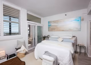 A bright and airy bedroom with a large bed dressed in white linens and multiple pillows. The room features white walls, a large window with white blinds, a glass door leading to an outdoor area, a small cushioned bench at the foot of the bed, a basket with a blanket, a small side table with decorative items, and a large beach-themed painting above the bed.