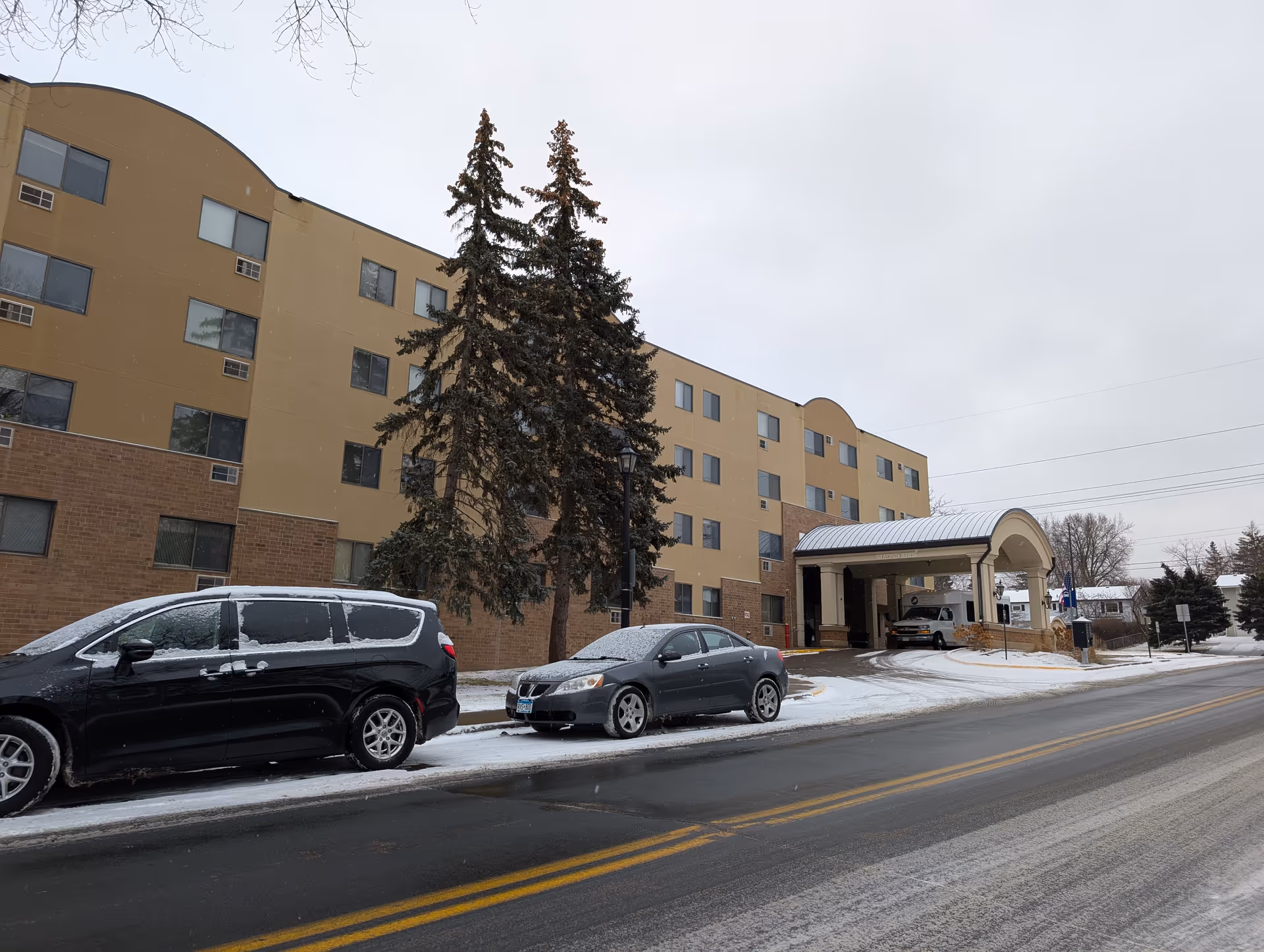 Front exterior of a multi-story senior living building with a covered entrance, parked cars, and light snow on the ground.